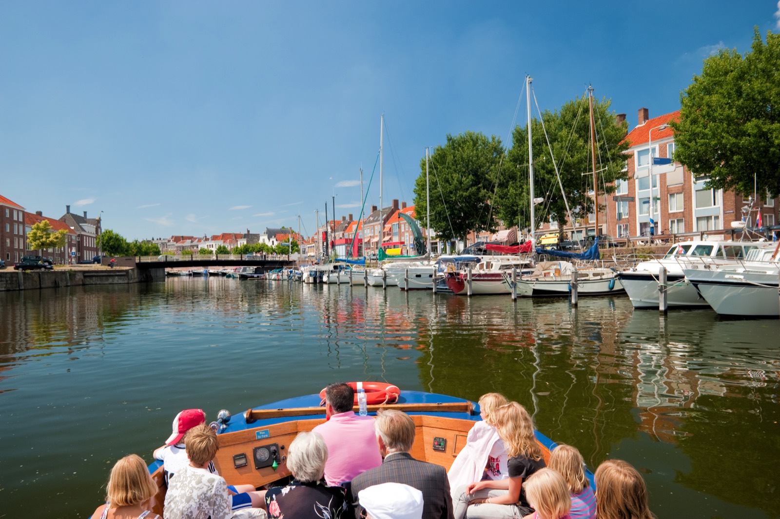 Gæster på agterdækket af en båd sejler ind i en havn med sejlbåde og rækkehuse